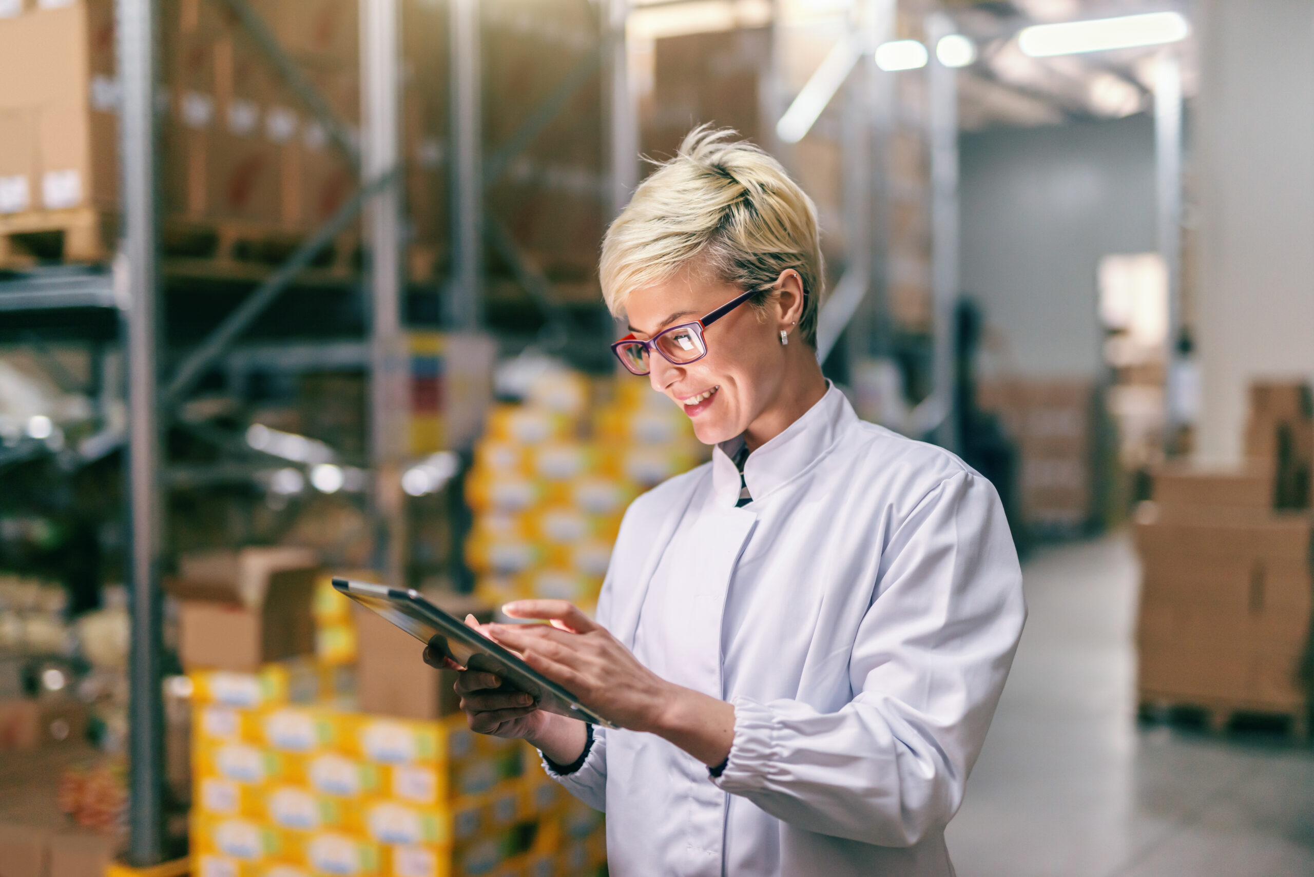 Young Caucasian blonde woman in white uniform using tablet in warehouse.
